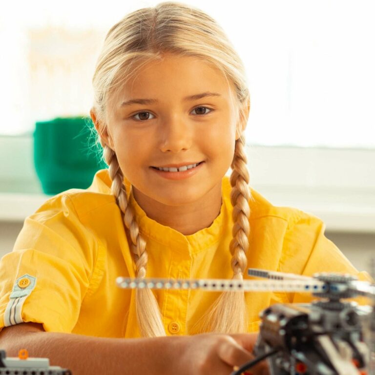 Cheerful blonde girl sitting at the desk with a helicopter model on it during her technology lesson